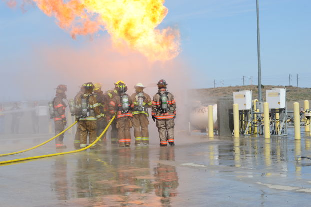 Eine Gruppe von Feuerwehrmännern in Helmen, die Rohre halten, steht vor einem großen Feuer mit verstreuten Pfählen, Drähten und anderen Gegenständen auf dem Boden unter einem bewölkten Himmel.