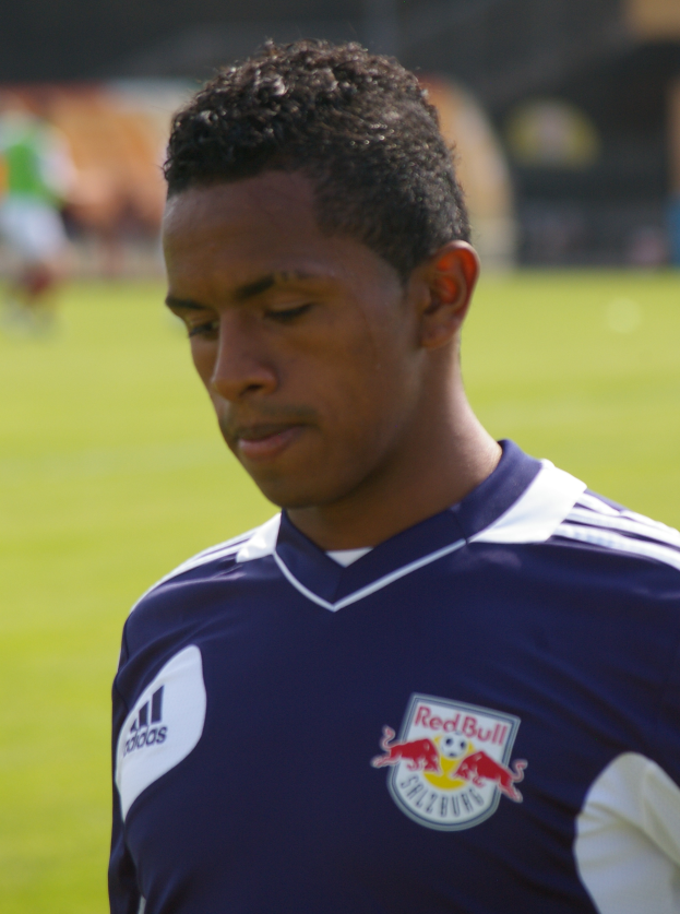 Ein junger Mann in einem blauen T-Shirt steht auf einem Fußballfeld, mit kurzen, dunklen Haaren und einem entschlossenen Gesichtsausdruck.