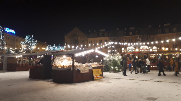 Eine verschneite Nachtansicht auf einem Weihnachtsmarkt mit Menschen, Buden mit Lichtern, Pflanzen, Bäumen, Gebäuden und Schildern unter einem bewölkten Himmel.