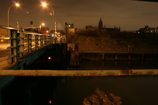 Eine Brücke überspannt einen Fluss mit mehreren Straßenlaternen auf ihrer Länge und zahlreichen Gebäuden im Hintergrund.