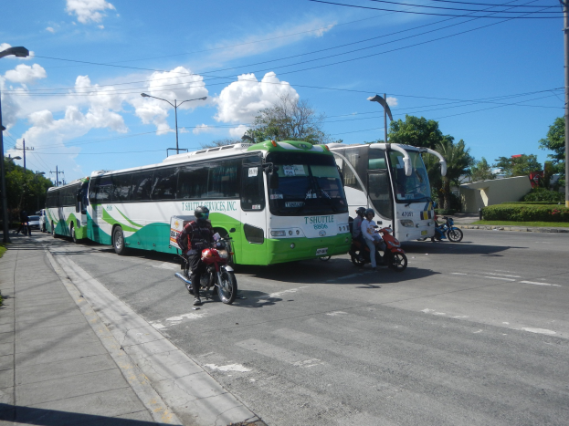 Ein grüner und weißer Shuttlebus steht am Straßenrand mit Motorradfahrern davor, ein grasbewachsener Fußweg links und Gebäude, Bäume und Laternen im Hintergrund unter einem klaren blauen Himmel.