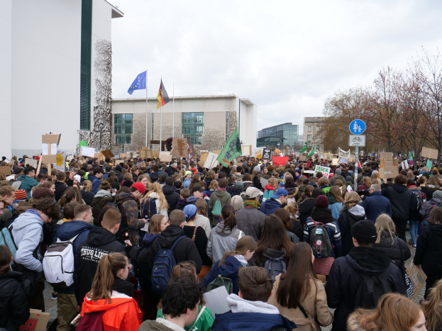 Eine große Gruppe von Menschen steht vor einem Gebäude und hält Schilder und Fahnen mit Stangen, wahrscheinlich bei einer Klimademonstration in Deutschland, mit Bäumen und Schildern im Hintergrund unter einem bewölkten Himmel.