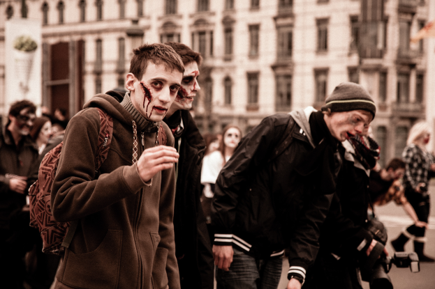 Eine Gruppe von Menschen in Zombie-Kostümen, einige in Kostümen, die eine Straße entlanggehen, mit einem Gebäude im Hintergrund, einer Kamera haltend.