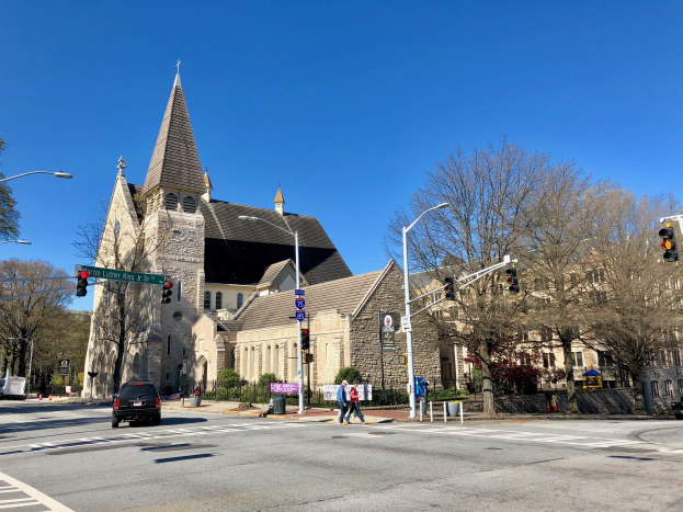 Die Episkopalkirche St. Lukas, eine große Kirche mit einem Turm, steht an einer Straßenecke umgeben von Gebäuden, Verkehr, Fußgängern und Grünflächen unter einem klaren blauen Himmel.