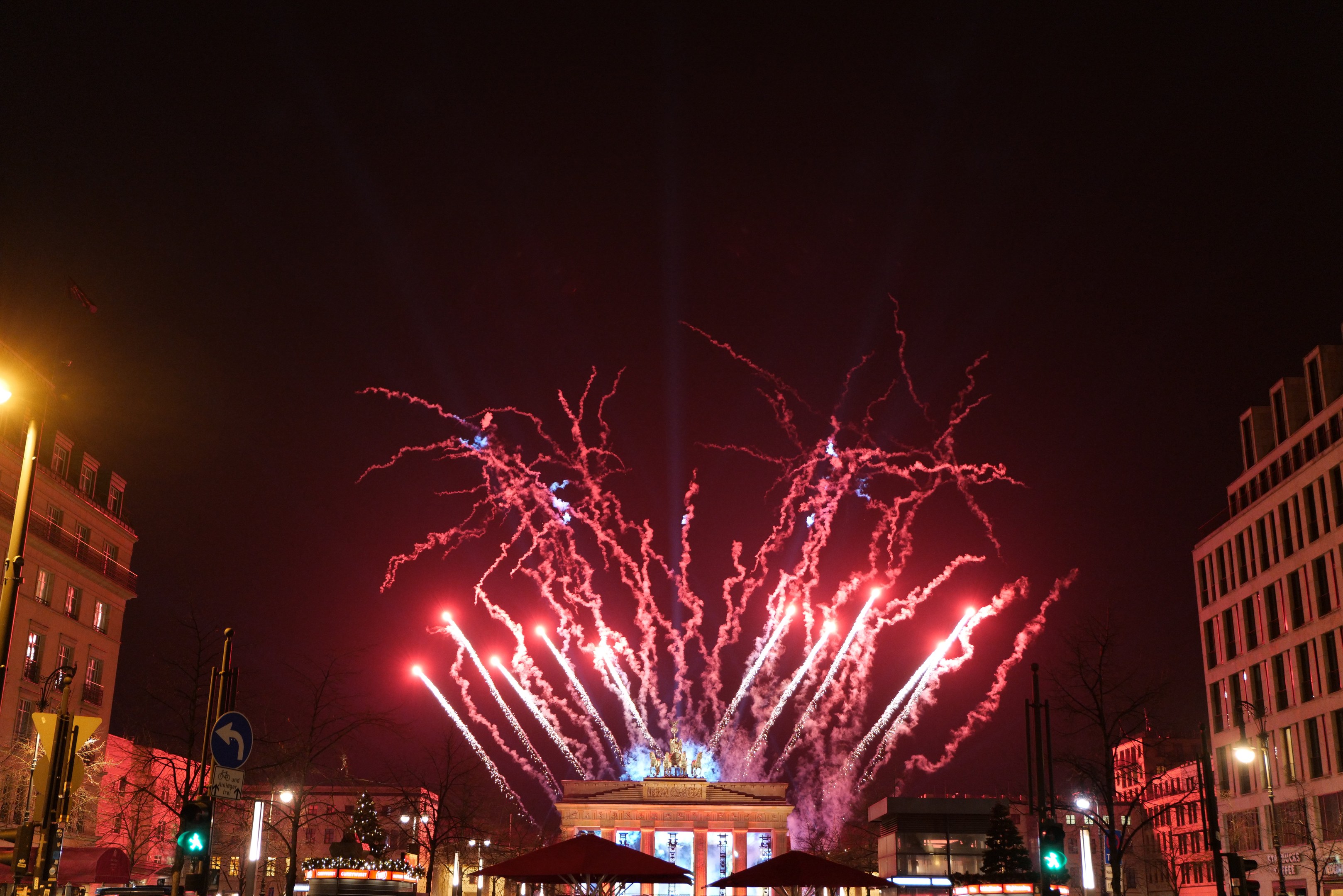 Eine belebte Stadtstraße in Berlin am Silvesterabend, voller Menschen, Fahrzeuge und festlicher Beleuchtung von Gebäuden und Feuerwerk am Himmel.