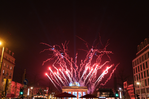Eine belebte Stadtstraße in Berlin am Silvesterabend, voller Menschen, Fahrzeuge und festlicher Beleuchtung von Gebäuden und Feuerwerk am Himmel.
