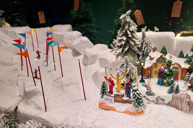 Eine festliche Weihnachtsdorfszene mit schneebedeckten Bäumen, Häusern, Menschen, Fahnen, Masten, Lichtern und Tieren vor einem dunklen, verschneiten Berg im Hintergrund.