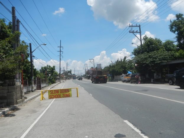 Eine Straße mit Fahrzeugen, ein "Verlangsamen Sie die Schule"-Schild, Strommasten mit Drähten, Bäume, Laternenmasten und ein bewölkter Himmel im Hintergrund.