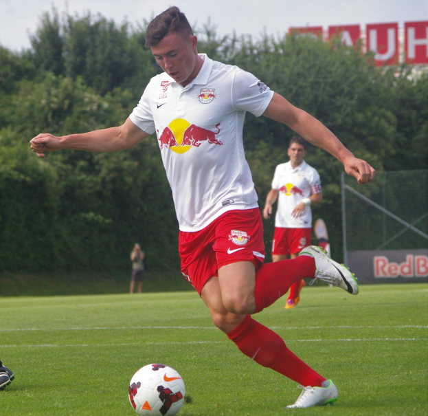 Ein Mann in einem Fu├čballtrikot schie├čt einen Ball auf einem gr├╝nen Feld mit B├Ąumen und einem klaren blauen Himmel im Hintergrund, mit einem Schild mit der Aufschrift "RB Leipzig" im Bild.