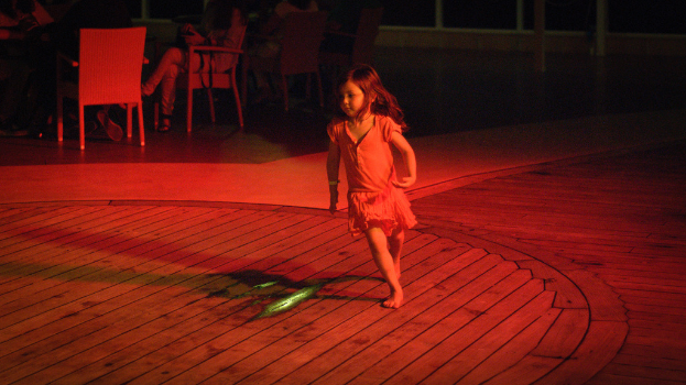 Ein Kind läuft über eine hölzerne Plattform drinnen, mit ein paar Menschen, die im Hintergrund auf Stühlen sitzen.