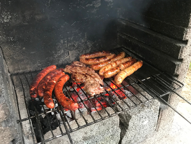 Grill mit Würstchen und Fleisch darauf, umgeben von brennender Kohle und Flammen, im Freien auf Gras gesetzt.