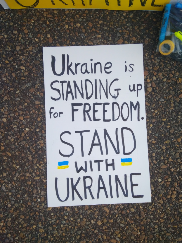 Ein weißes Blatt Papier mit dem Text "Ukraine steht für die Freiheit ein, steht mit Ukraine" vor einer gelben Fahne auf dem Boden. Rechts davon ein Plastikbehälter mit verschiedenen Gegenständen.