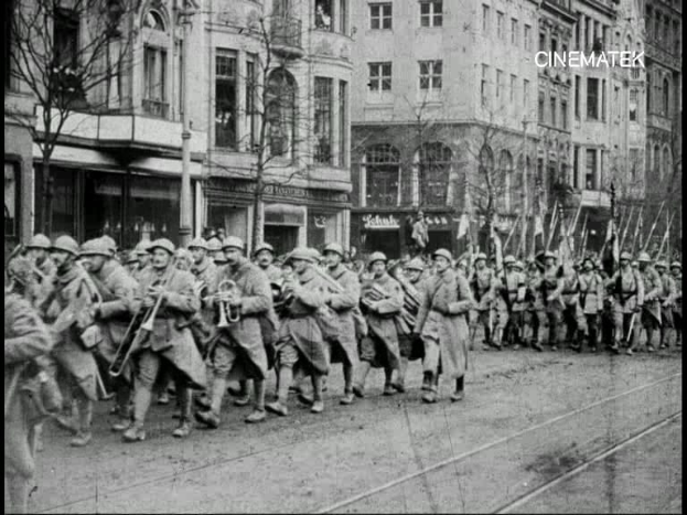Schwarzes und weißes Foto von Soldaten, die während des Ersten Weltkriegs die Straße in Berlin entlangmarschieren, mit Mützen und Gewehren, sowie Bäumen und Gebäuden an der Straße und einem Pfahl im Hintergrund.