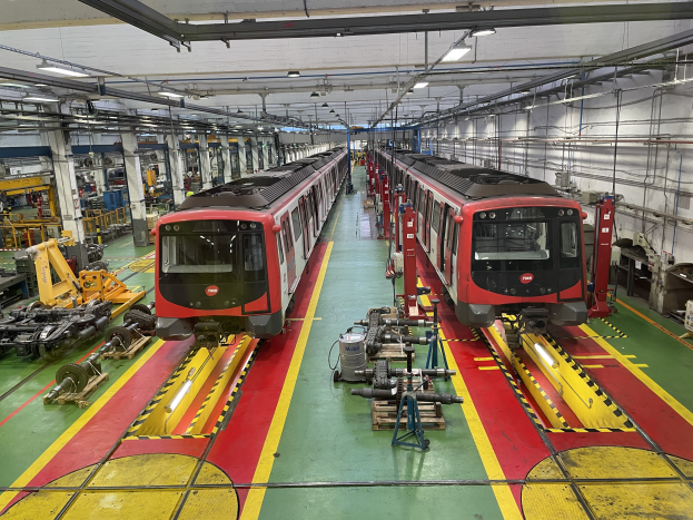 Zwei Züge auf Schienen innerhalb einer großen Industriehalle mit Maschinen und Geräten, mit einer Wand mit Rohren und Deckenleuchten auf der rechten Seite, betrieben von der Daimler AG.