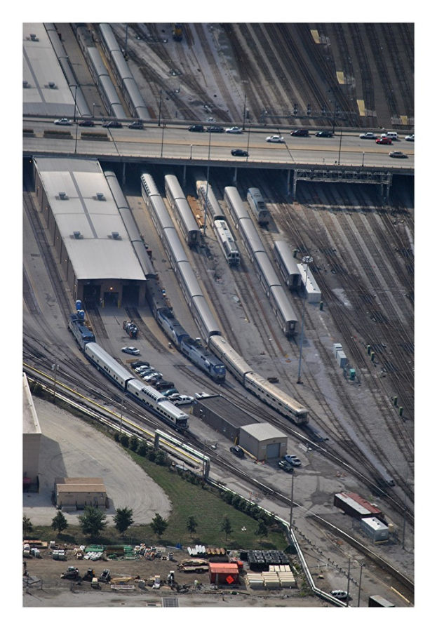 Eine Brücke mit einigen Fahrzeugen darauf, darunter Eisenbahnschienen mit einigen Zügen und eine Landschaft mit Gebäuden, Bäumen, Pfählen und verschiedenen Objekten und Fahrzeugen auf dem Boden.
