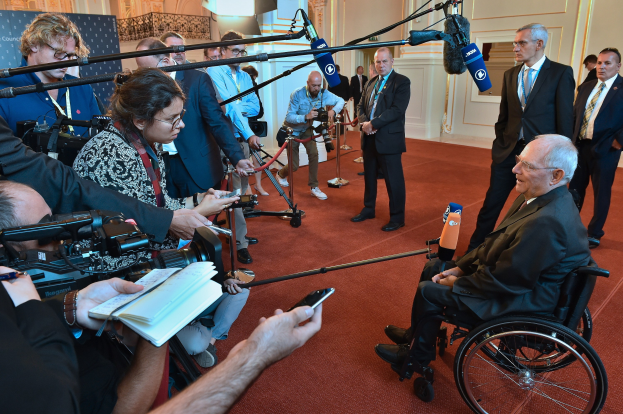 Ein Mann im Anzug im Rollstuhl spricht zu Reportern mit Kameras und Mikrofonen, mit einem Banner, einer Wand und einer Tür im Hintergrund.