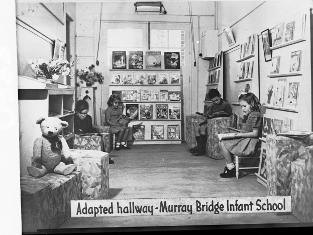 Gruppe von Kindern auf Stühlen in einem bibliotheksähnlichen Raum mit Bücherregalen, einem Teddybär links, einer Blumenvase auf dem Tisch, gerahmten Gegenständen an der Wand und Text unten lesend "Angepasster Flur Murray Bridge Kindergarten"