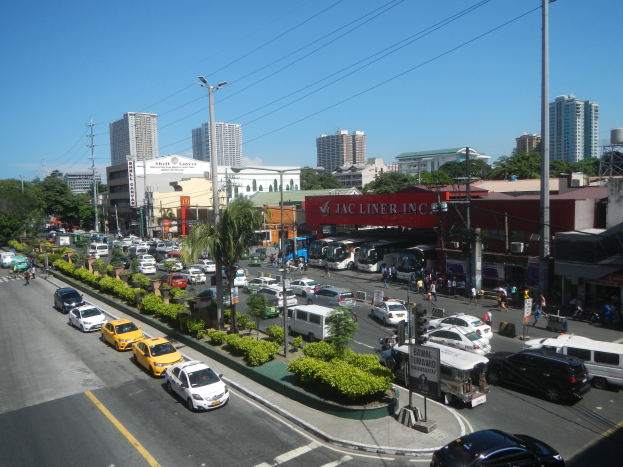 Eine belebte Stadtstraße mit zahlreichen Autos, Bussen und Fußgängern, umgeben von Gebäuden, Bäumen und verschiedener städtischer Infrastruktur unter einem klaren blauen Himmel.