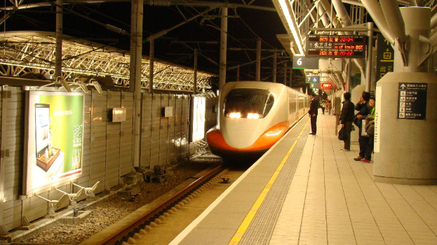 Eine Gruppe von Menschen steht auf einem Bahnsteig mit einem Zug auf den Schienen, eine Wand mit einer Tafel links und mehrere Tafeln an Pfählen rechts.