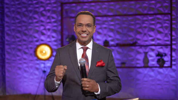 Stephen Colbert, mit Anzug, Krawatte und Brille, steht auf einer B├╝hne und h├Ąlt ein Mikrofon, mit einer Wand und verschiedenen Gegenst├Ąnden dahinter.