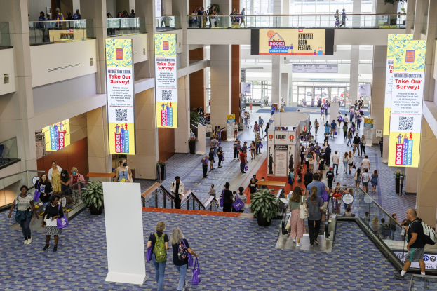 Eine große Menschenmenge bewegt sich durch ein großes Gebäude mit Bannern, Topfpflanzen, Rolltreppen, Geländern, Säulen, Texttafeln, Glastüren und anderen Elementen, einige tragen Taschen, was auf die Teilnahme an der Nationalen Buchmesse hinweist.