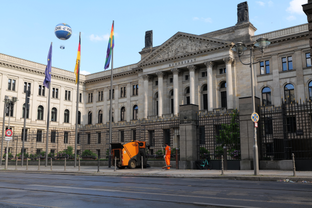 Das Reichstagsgebäude in Berlin, Deutschland, ein großes Gebäude mit zahlreichen Fenstern, umgeben von Straßenmasten, Laternen, Schildern, Fahrzeugen, Fußgängern, Flaggen, Gittern und Bäumen unter einem bewölkten Himmel.