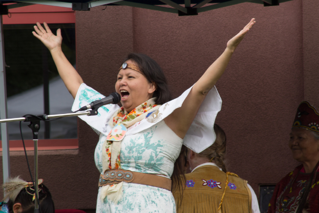 Eine Frau in einem weißen Kleid steht auf einer Bühne und spricht in ein Mikrofon, mit einer Wand und einem Eisenrahmen dahinter.