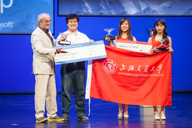 Vier Personen auf einer Bühne mit Auszeichnungen, Schecks und einer Fahne, strahlend, mit einem digitalen Bildschirm im Hintergrund.
