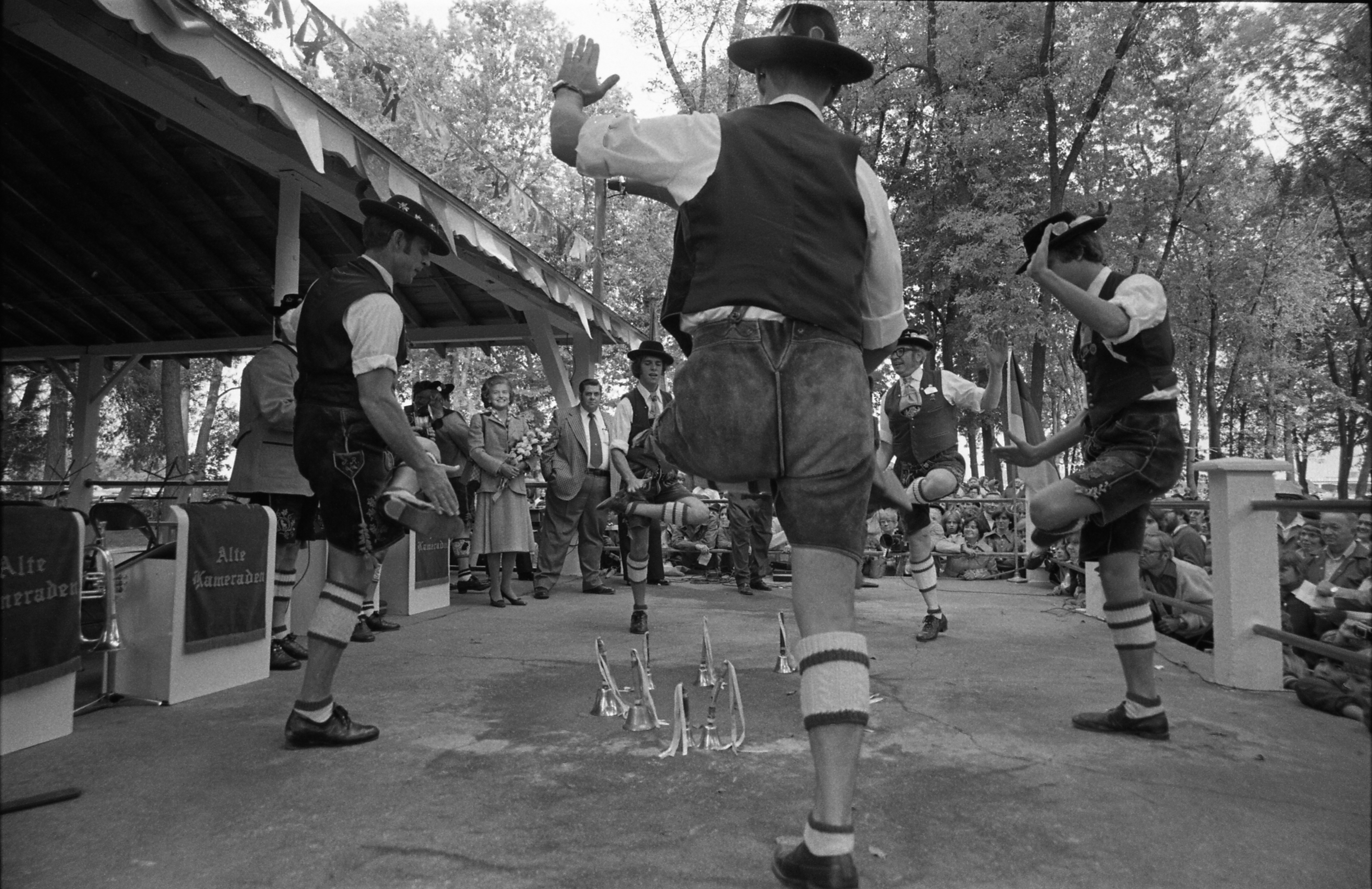 Eine Gruppe von Männern in traditioneller bayrischer Tracht tanzt auf einer Bühne vor einem gemischten Publikum, mit Bäumen und einem Schuppen im Hintergrund, dargestellt in Schwarz-Weiß.