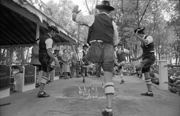 Eine Gruppe von Männern in traditioneller bayrischer Tracht tanzt auf einer Bühne vor einem gemischten Publikum, mit Bäumen und einem Schuppen im Hintergrund, dargestellt in Schwarz-Weiß.