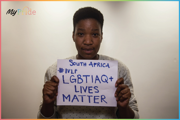 Eine Frau in einem weißen Hemd hält entschlossen ein Schild mit der Aufschrift "LGBTQ+ Lives Matter" vor einer Wand.