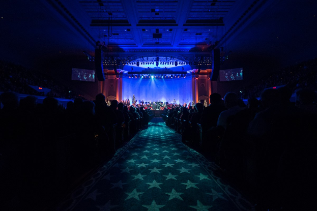 Großer Zuschauerraum mit Sitzplätzen, Bühne mit Musikern, Deckenleuchten und Fernsehern, dunkle Konzerthallenatmosphäre.