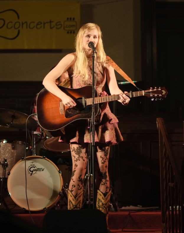 Eine Frau spielt Gitarre und singt auf der Bühne eines Konzerts, mit einer Band und einem Bildschirm im Hintergrund und einer Bühne unten.