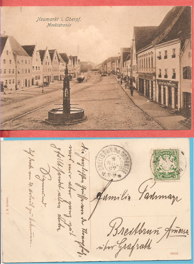 Eine alte Postkarte, die eine Stadtstraße mit Gebäuden, Straßen, Fahrzeugen, Menschen und einem Brunnen zeigt und oben einen Stempel und unten Text hat.