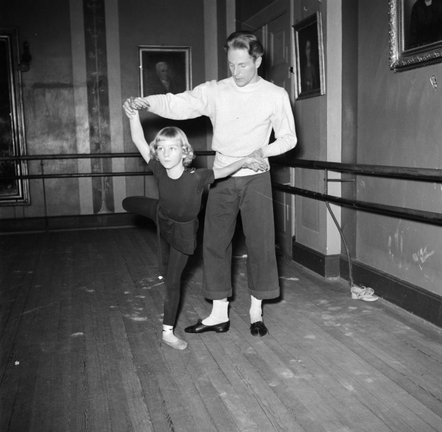 Ein Mann in einem weißen Hemd und schwarzen Hosen tanzt mit einem kleinen Mädchen in einem Tanzstudio, mit Fotoframes an den Wänden und einem Geländer im Hintergrund, alles in Schwarz-Weiß dargestellt.