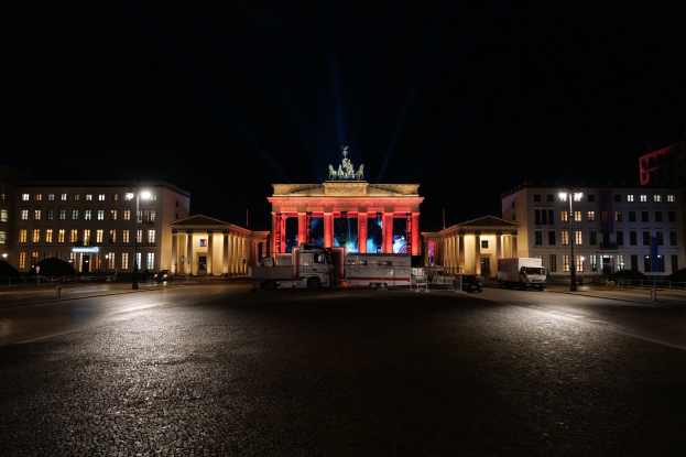 Das Brandenburger Tor in Berlin bei Nacht, beleuchtet von Lichtern, mit Fahrzeugen auf der Straße und Gebäuden mit Fenstern, Laternenmasten und einer Statue im Hintergrund unter einem dunklen Himmel.