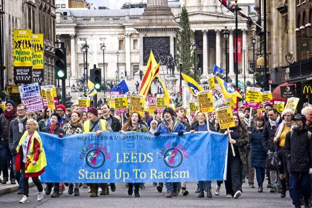 Eine Gruppe von Menschen marschiert mit einer 'Stand Up to Racism'-Schleife die Straße entlang, mit Gebäuden, Bäumen, Laternen, Ampeln und einer Statue im Hintergrund unter einem bewölkten Himmel.