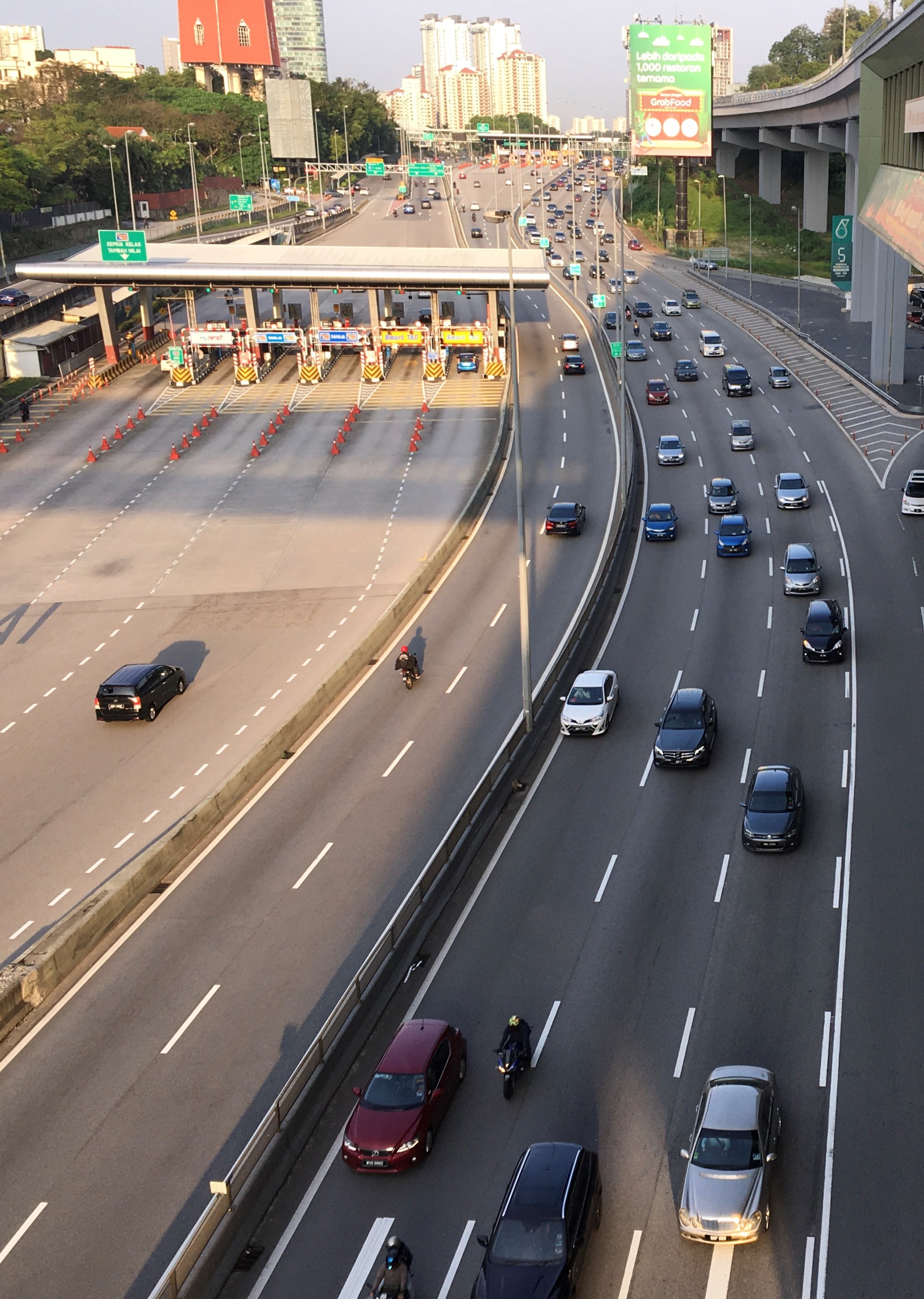 Eine belebte Autobahn mit zahlreichen Autos, Motorrädern, Verkehrskegeln, Verkehrszeichen, Laternenmasten, einer Brücke, Bäumen, Gebäuden unter einem klaren blauen Himmel.
