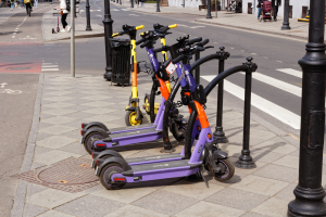 Eine Reihe von Elektro-Scootern, die auf der Straße geparkt sind, mit Menschen, die auf dem Gehweg laufen und Fahrräder fahren, ein Müllcontainer im Vordergrund und Gebäude, Bäume und Pflanzen im Hintergrund.