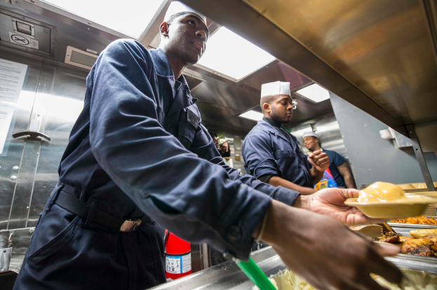 Eine Gruppe von Männern in einer Küche, einer mit einem Teller Essen haltend, mit Tabletts voller Essen auf dem Tisch, einem Feuerlöscher und Papieren an der Wand im Hintergrund.