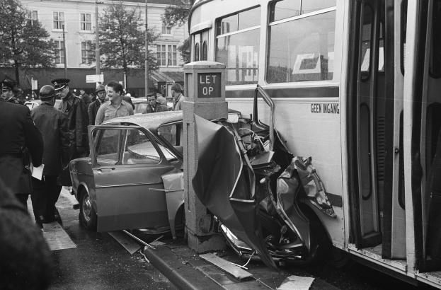 Schwarzes und weißes Bild eines Busses, der auf der Straße in ein Auto gekracht ist, mit Menschen um die Unfallstelle und Bäumen und Gebäuden im Hintergrund.
