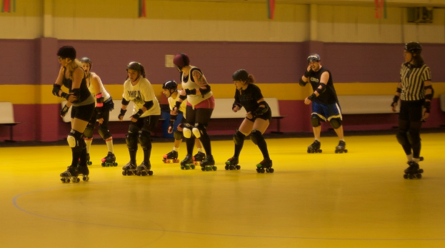 Leute in Kleidung sind auf einem Boden mit Bänken vor einer Wand Schlittschuh fahrend.