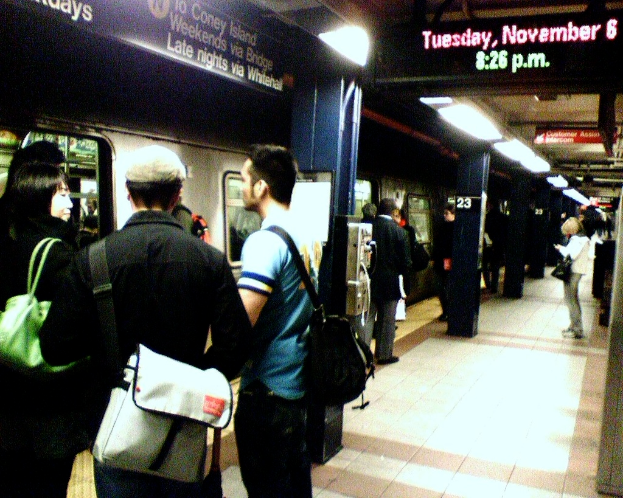 Ein überfüllter Bahnsteig mit Menschen mit Taschen, ein Zug links und ein Textanzeigebrett oben.