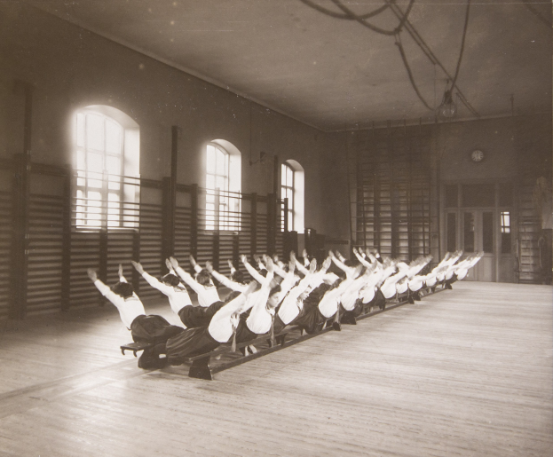 Gruppe von Menschen, die Pilates-Übungen in einem Fitnessstudio mit Fenstern und einer Uhr an der Wand im Hintergrund durchführt.