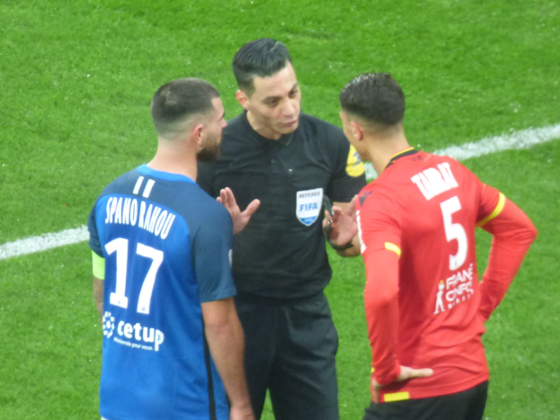 Schiedsrichter in schwarzem Hemd und Hose, der mit zwei Männern auf einem Fußballfeld spricht, einer in blau-roten T-Shirts mit Text, beide tragen Brillen.