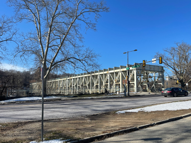 Ein Auto fährt auf einer schneebedeckten Straße neben einer Brücke entlang, mit Laternen, Ampeln, Bäumen und einem bewölkten Himmel im Hintergrund.