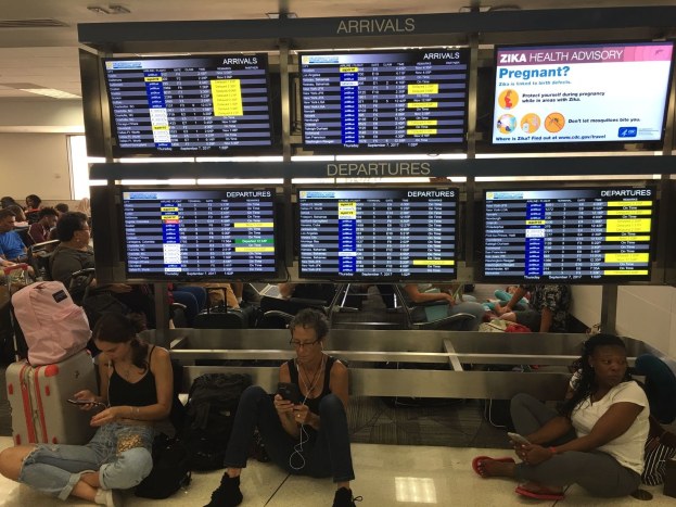 Eine Gruppe von Menschen sitzt auf dem Boden eines Flughafens, einige halten Handys, mit verstreuten Taschen und Zika-Virus-bezogenen Bildschirmen im Hintergrund.