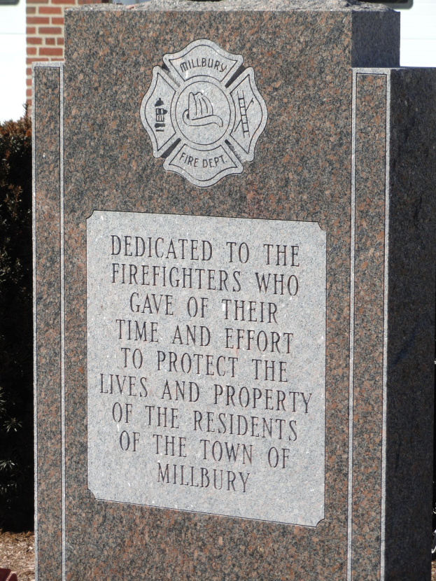 Gedenkstein mit einer Plakette für Feuerwehrleute in Millbury, vor einem Backsteingebäude inmitten von Grünanlagen.