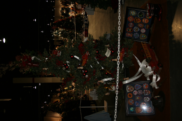 Ein Weihnachtsbaum mit Geschenken, Rahmen und Spielzeugen auf einer Plattform, mit Menschen, zusätzlichen Weihnachtsbäumen, Ketten, Pfählen und Lichtern im Hintergrund.