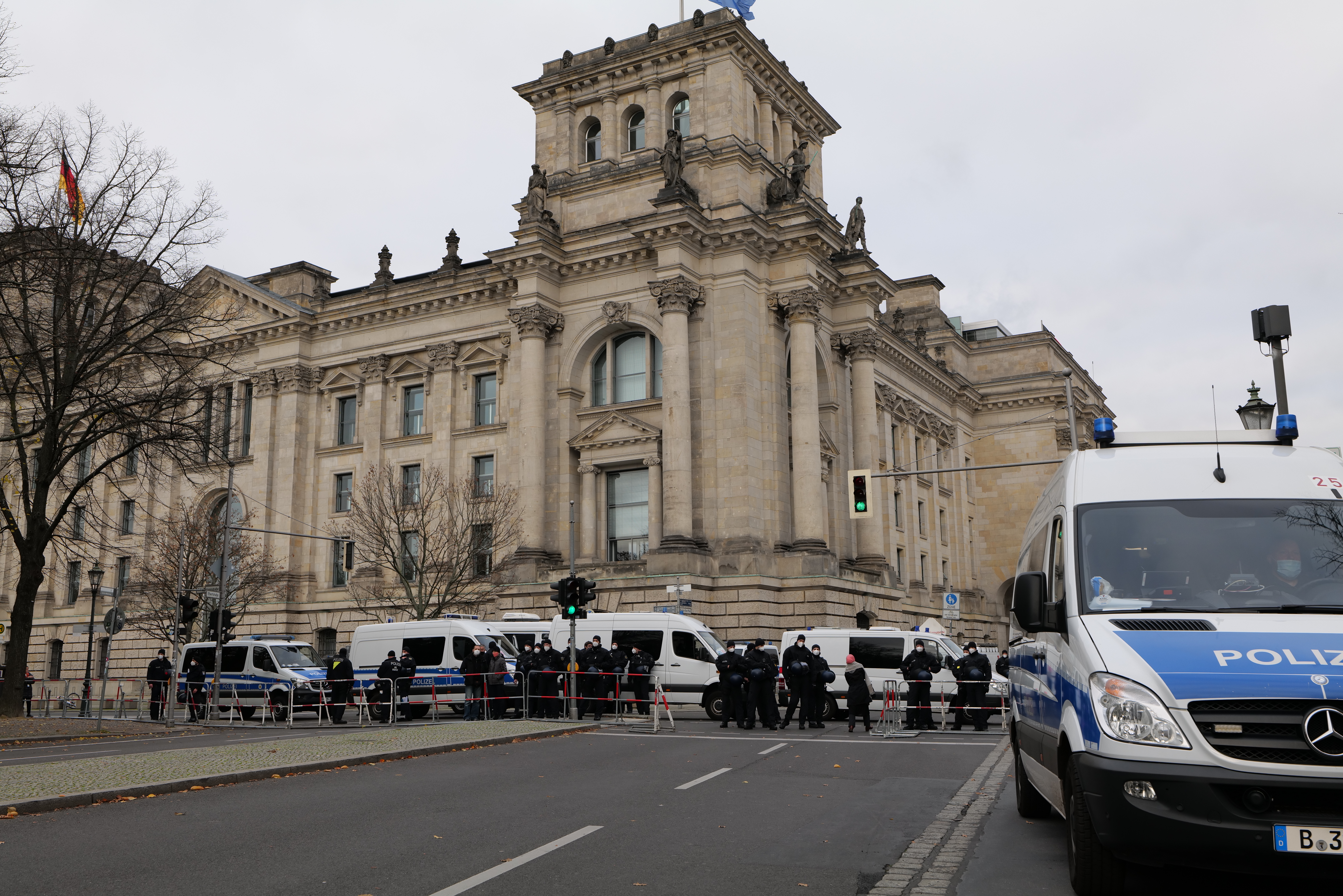 Eine Gruppe von Polizisten steht vor dem Reichstaggebäude in Berlin, Deutschland, mit Fahrzeugen, einem Zaun, Ampeln, Laternenpfählen, Bäumen und Fahnen im Hintergrund, unter einem klaren Himmel.